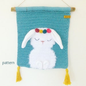 May include: A crocheted wall hanging featuring a white bunny with a colourful flower crown. The bunny is sitting on a teal blue crocheted background. The wall hanging has two yellow tassels.
