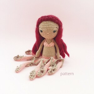 May include: Crocheted mermaid doll with red hair and a pink tail. The doll is sitting on a pink background. pattern