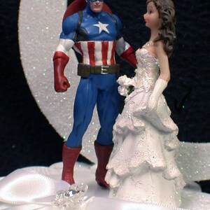 May include: A wedding cake topper featuring a superhero in a blue and red costume with a star on his chest and a white bride in a white gown. The bride is holding a bouquet of white flowers.
