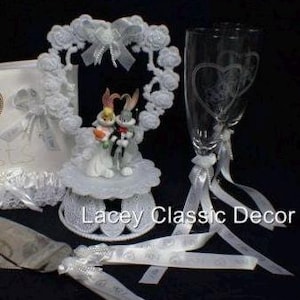 Puede incluir: Un conjunto de decoración de boda con figuras de Bugs Bunny y Lola Bunny, un arco floral blanco y copas de champán con diseños de corazón. El conjunto incluye un servidor de pastel y una liga, todo adornado con cintas blancas.