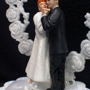 May include: A white wedding cake topper featuring a bride and groom in a dancing pose. The bride is wearing a white dress with a sparkly bodice and the groom is wearing a black tuxedo. The cake topper is set on a white cake base with a white satin ribbon and a sparkly rhinestone.