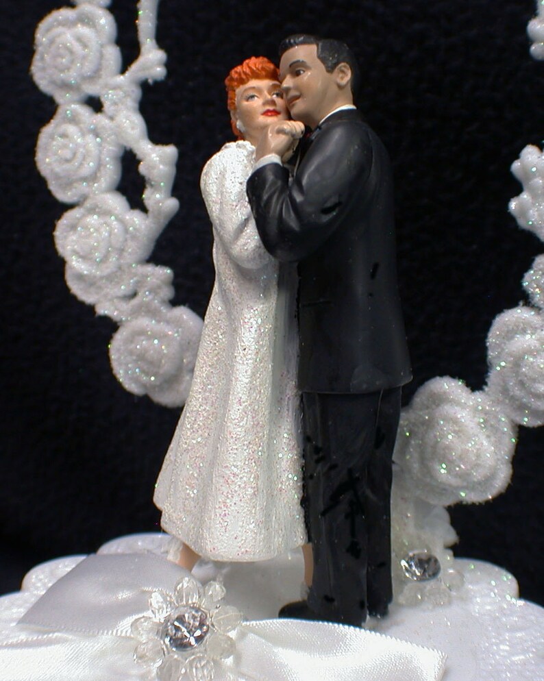 May include: A white wedding cake topper featuring a bride and groom in a dancing pose. The bride is wearing a white dress with a sparkly bodice and the groom is wearing a black tuxedo. The cake topper is set on a white cake base with a white satin ribbon and a sparkly rhinestone.