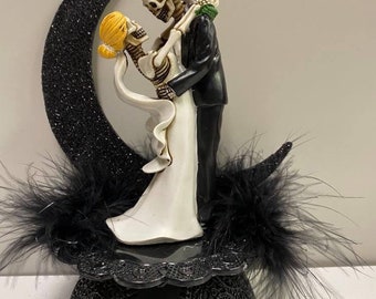 Op de afbeelding: Een Halloween-thema bruidstaarttopper met een omhelzend skeletbruidspaar. De bruidegom draagt een zwart smoking, de bruid een witte jurk. Een zwarte glittermaan en een decoratieve basis met zwarte veren maken het ontwerp compleet.