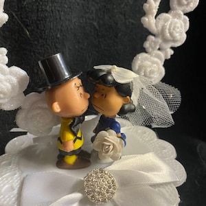 以下が含まれることがあります： 白いケーキトッパーに、結婚式用の衣装を着たチャーリーブラウンとスヌーピーの漫画のカップルが描かれています。カップルは、白いリボンとラインストーンの装飾が施された白い台の上に立っています。