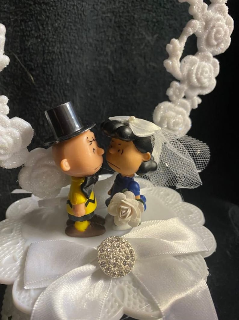 以下が含まれることがあります： 白いケーキトッパーに、結婚式用の衣装を着たチャーリーブラウンとスヌーピーの漫画のカップルが描かれています。カップルは、白いリボンとラインストーンの装飾が施された白い台の上に立っています。