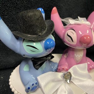 May include: A cake topper featuring two cartoon character figurines, one blue and one pink. The blue character wears a black top hat and bow tie. The pink character has a white bow. The topper sits on a white, scalloped base.