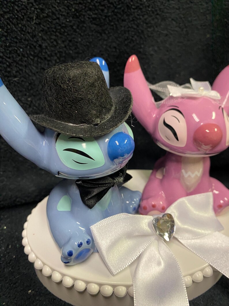 May include: A cake topper featuring two cartoon character figurines, one blue and one pink. The blue character wears a black top hat and bow tie. The pink character has a white bow. The topper sits on a white, scalloped base.
