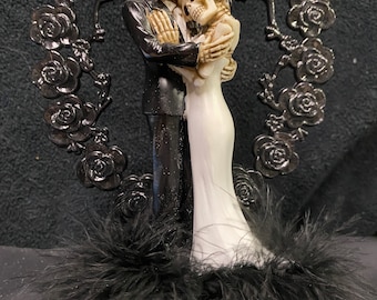 May include: A black and white skeleton wedding cake topper with a heart-shaped rose design. The skeletons are holding each other and are dressed in a tuxedo and a white wedding dress.