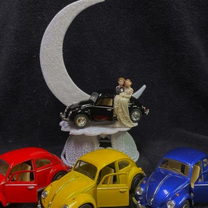 Puede incluir: Un adorno para pastel de boda con una figura de novia y novio sentados en un coche negro, con una luna creciente blanca arriba. Tres coches de juguete Volkswagen Beetle se muestran debajo, uno rojo, uno amarillo y uno azul.