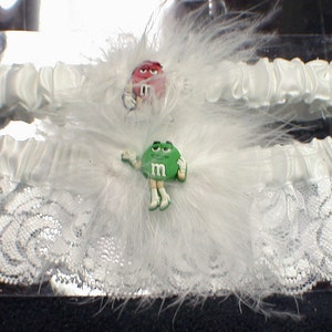 May include: Two white satin garters with white feathers. One garter has a red M&M's candy character and the other has a green M&M's candy character.
