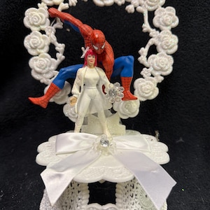 May include: A cake topper featuring a Spider-Man figure crouching behind a figure in a white outfit, set against a white glittery rose-shaped frame. The base is a white scalloped platform decorated with a white satin ribbon and a central rhinestone.