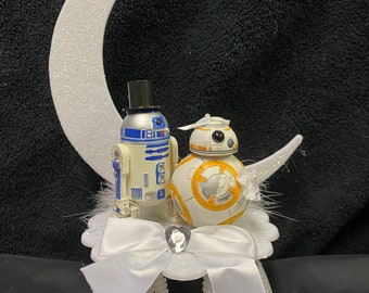 May include: A white cake topper featuring a white crescent moon with glitter, a white and blue R2-D2 figurine wearing a black top hat, and a white and orange BB-8 figurine wearing a white veil. The figurines are standing on a white cake stand with a white bow and a rhinestone.