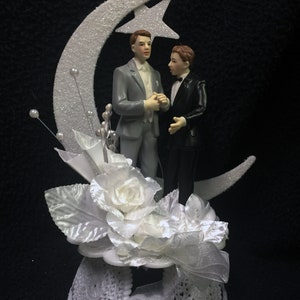 May include: A wedding cake topper featuring two grooms in formal attire. They stand before a glittery crescent moon and star. The base includes white flowers, leaves, and decorative elements, all in white.