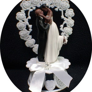 Puede incluir: Topper de pastel de boda en blanco y negro con una novia y un novio abraz&aacute;ndose. La novia lleva un vestido blanco y el novio lleva un esmoquin negro. La pareja est&aacute; de pie frente a un dise&ntilde;o floral en forma de coraz&oacute;n blanco.
