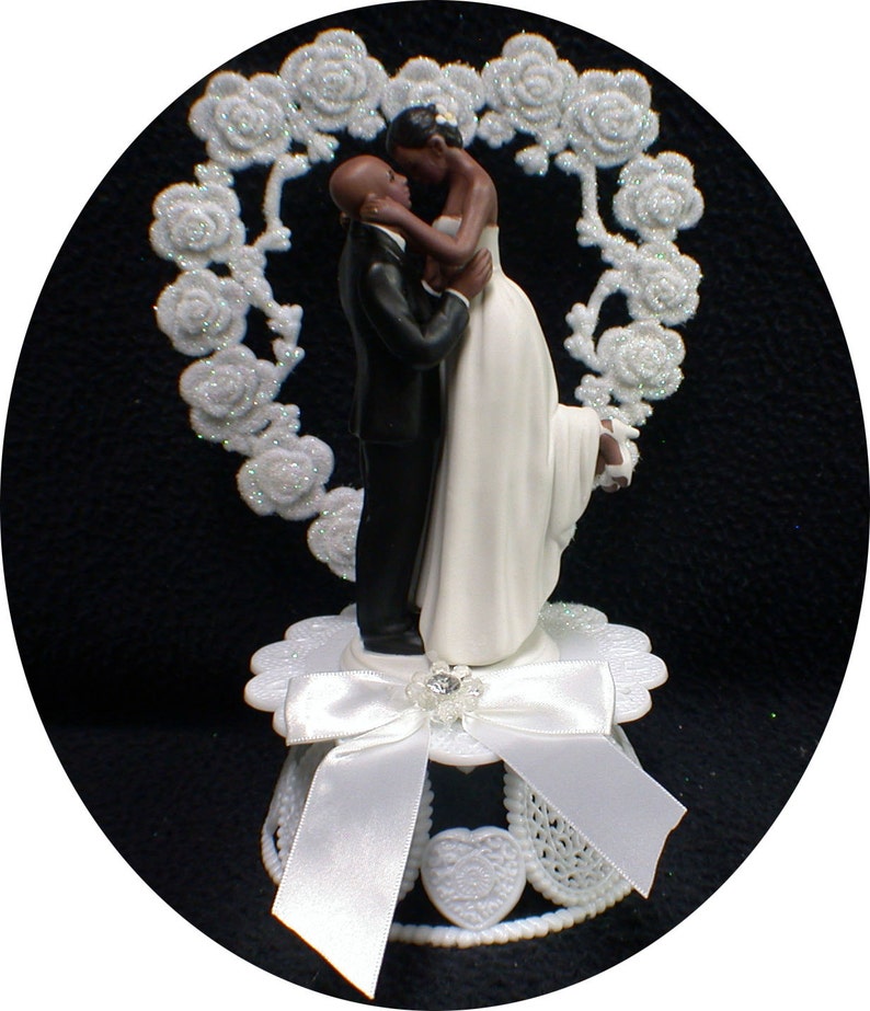 Puede incluir: Topper de pastel de boda en blanco y negro con una novia y un novio abraz&aacute;ndose. La novia lleva un vestido blanco y el novio lleva un esmoquin negro. La pareja est&aacute; de pie frente a un dise&ntilde;o floral en forma de coraz&oacute;n blanco.
