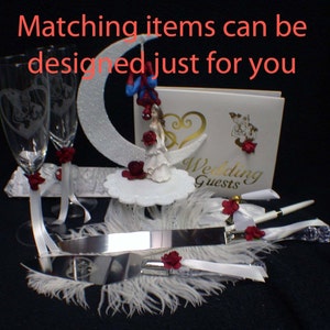May include: Wedding cake topper with a bride and groom figurine, a white cake knife, two champagne flutes, a guest book, and a white feather boa. The cake topper features a bride and groom figurine standing on a crescent moon. The bride is wearing a white dress and the groom is wearing a blue suit. The cake knife is silver and has a white handle. The champagne flutes are clear glass and have a white ribbon tied around the stem. The guest book is white and has the words "Wedding Guests" printed on the front. The white feather boa is draped over the cake knife and champagne flutes.
