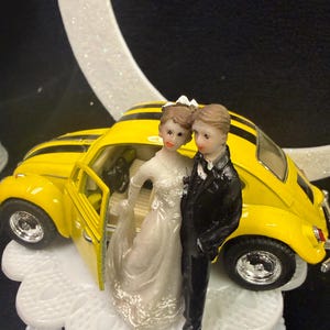 Puede incluir: Un adorno para pastel que presenta un coche miniatura amarillo con rayas negras y dos figuras de pie frente a él. Una figura lleva un vestido blanco y la otra un traje negro. Todo el conjunto está montado sobre una base blanca festoneada.