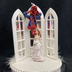 Può includere: Un topper per torta nuziale bianco con una statuina di sposa e sposo. Lo sposo è vestito da Spiderman, appeso a testa in giù da una cornice di finestra bianca, tenendo un mazzo di rose rosse. La sposa indossa un abito bianco e tiene un mazzo di fiori bianchi.