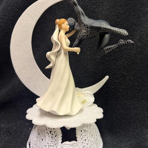 May include: A wedding cake topper featuring a bride in a white gown standing before a large, glittery white crescent moon. A black spider-themed figure is suspended from the top of the moon, reaching toward the bride. The base is a white, lacy stand.