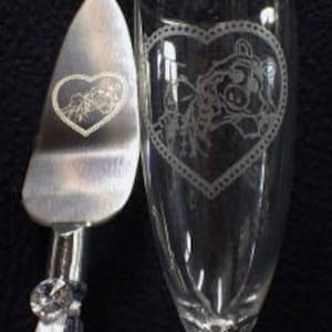 Puede incluir: Un servidor de pastel de plata y una flauta de champ&aacute;n con un dise&ntilde;o grabado de una pareja que se toma de la mano dentro de un coraz&oacute;n. El dise&ntilde;o est&aacute; rodeado por un borde decorativo.