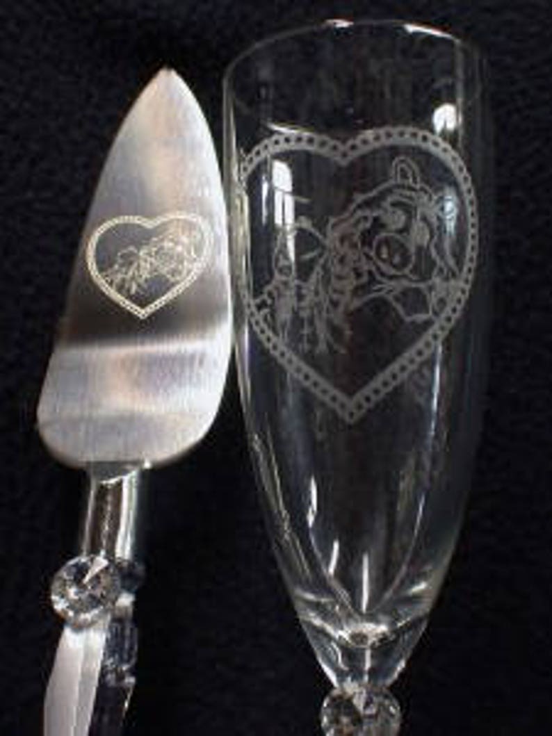 Puede incluir: Un servidor de pastel de plata y una flauta de champ&aacute;n con un dise&ntilde;o grabado de una pareja que se toma de la mano dentro de un coraz&oacute;n. El dise&ntilde;o est&aacute; rodeado por un borde decorativo.