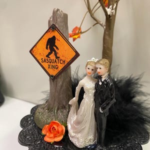 Puede incluir: Un adorno para pastel de bodas con una figura de novia y novio. La novia lleva un vestido blanco y el novio un esmoquin negro. Un letrero que dice "Sasquatch Xing" está al lado de un árbol con hojas naranjas. Una base negra con una rosa.