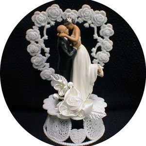 May include: A white cake topper featuring a bride and groom in a heart-shaped frame. The bride is wearing a white dress and the groom is wearing a black suit. The couple is kissing.