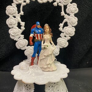May include: White cake topper featuring a bride and groom figurine. The bride is wearing a white dress and the groom is wearing a blue and red costume. The topper is decorated with white roses and a heart-shaped frame.
