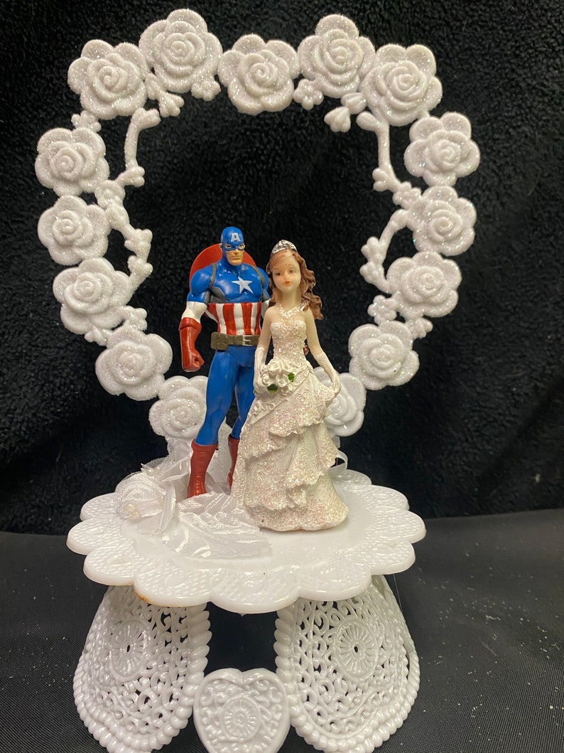 May include: White cake topper featuring a bride and groom figurine. The bride is wearing a white dress and the groom is wearing a blue and red costume. The topper is decorated with white roses and a heart-shaped frame.