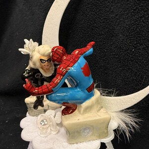 May include: A ceramic figurine featuring Spider-Man in a red and blue suit and Black Cat in a black and white suit, both perched on a stone-like base. Behind them is a large, glittery white crescent moon, all set atop a white lace-patterned stand.
