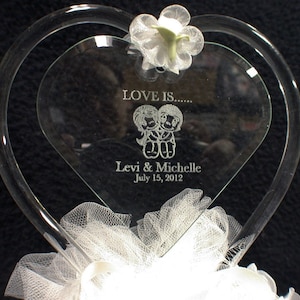 May include: A clear glass heart-shaped cake topper with a white tulle bow and a white flower. The heart is engraved with the text "LOVE IS..." and a cartoon couple, "Levi & Michelle" and "July 15, 2012".