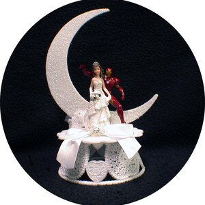 May include: White wedding cake topper featuring a bride and groom figurine standing on a crescent moon. The bride is wearing a white dress and the groom is wearing a red and gold suit. The base of the topper is white with a pearl trim.