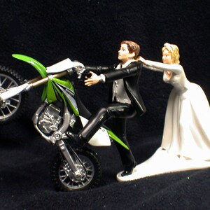 May include: A wedding cake topper featuring a groom in a tuxedo riding a green and white dirt bike on one wheel, while the bride in a white wedding dress pulls him back.