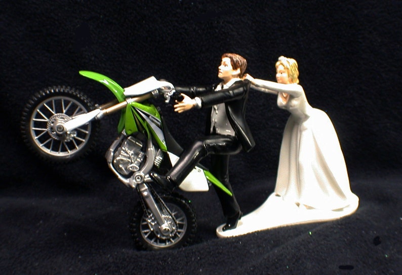 May include: A wedding cake topper featuring a groom in a tuxedo riding a green and white dirt bike on one wheel, while the bride in a white wedding dress pulls him back.