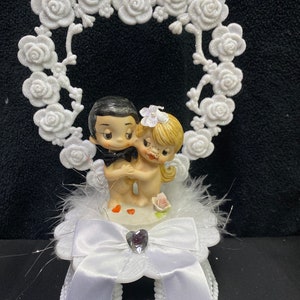 May include: A cake topper featuring a cartoon-style couple figurine standing on a white base decorated with white feathers, a white satin ribbon bow, and a small heart-shaped gem. Behind the couple is a large, glittery white rose-shaped arch.