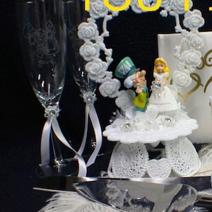 Könnte beinhalten: Weißer Hochzeitstortenaufsatz mit Alice im Wunderland Figuren, dem Hutmacher und Alice, mit einem weißen Rosenherz und weißem Band. Der Aufsatz ist auf einer weißen Torte mit einer weißen Feder und einem silbernen Tortenmesser.