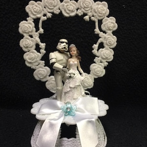 Może przedstawiać: Biały topper na tort ślubny przedstawiający figurkę szturmowca z Gwiezdnych Wojen trzymającego za rękę figurkę panny młodej. Para stoi pod białym, sercowatym łukiem kwiatowym. Podstawa topera jest biała z białą satynową kokardką i niebieskim kwiatem.