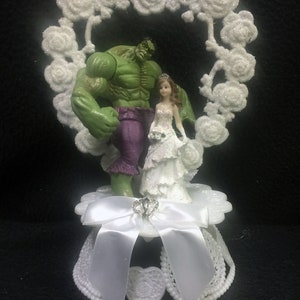 May include: A white cake topper featuring a green Hulk figurine and a white bride figurine standing under a white heart-shaped floral arch. The base of the topper is white with a white satin bow and a silver ring.