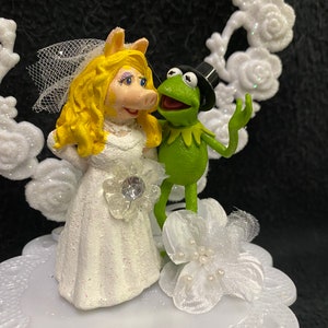 Puede incluir: Un adorno de pastel de bodas blanco con Miss Piggy con un vestido de novia blanco y Kermit la Rana con un traje verde y un sombrero de copa negro.