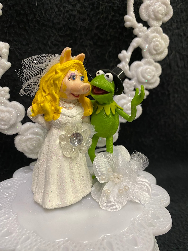 Puede incluir: Un adorno de pastel de bodas blanco con Miss Piggy con un vestido de novia blanco y Kermit la Rana con un traje verde y un sombrero de copa negro.