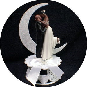 Puede incluir: Topper de pastel de bodas en blanco y negro con una novia y un novio bes&aacute;ndose en una luna creciente. La novia lleva un vestido blanco y el novio lleva un traje negro. La base del topper est&aacute; decorada con un lazo blanco y encaje.