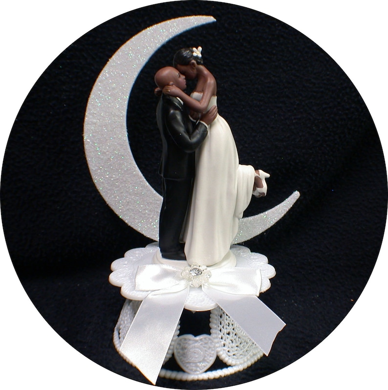 Puede incluir: Topper de pastel de bodas en blanco y negro con una novia y un novio bes&aacute;ndose en una luna creciente. La novia lleva un vestido blanco y el novio lleva un traje negro. La base del topper est&aacute; decorada con un lazo blanco y encaje.