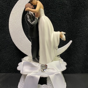 Pu&ograve; includere: Un topper per torta bianco con una sposa e uno sposo che si abbracciano sotto una luna crescente. La sposa indossa un abito bianco e lo sposo indossa uno smoking nero. La base del topper &egrave; decorata con un fiocco bianco e un strass.