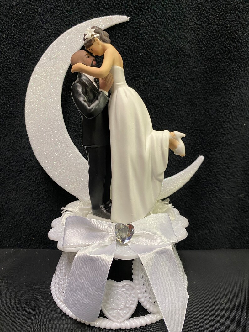 Pu&ograve; includere: Un topper per torta bianco con una sposa e uno sposo che si abbracciano sotto una luna crescente. La sposa indossa un abito bianco e lo sposo indossa uno smoking nero. La base del topper &egrave; decorata con un fiocco bianco e un strass.
