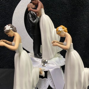 Pu&ograve; includere: Un topper per torta nuziale con una coppia che si bacia sotto una luna crescente, con altre due figure inginocchiate su entrambi i lati. Le figure sono tutte vestite con abiti da sposa bianchi.