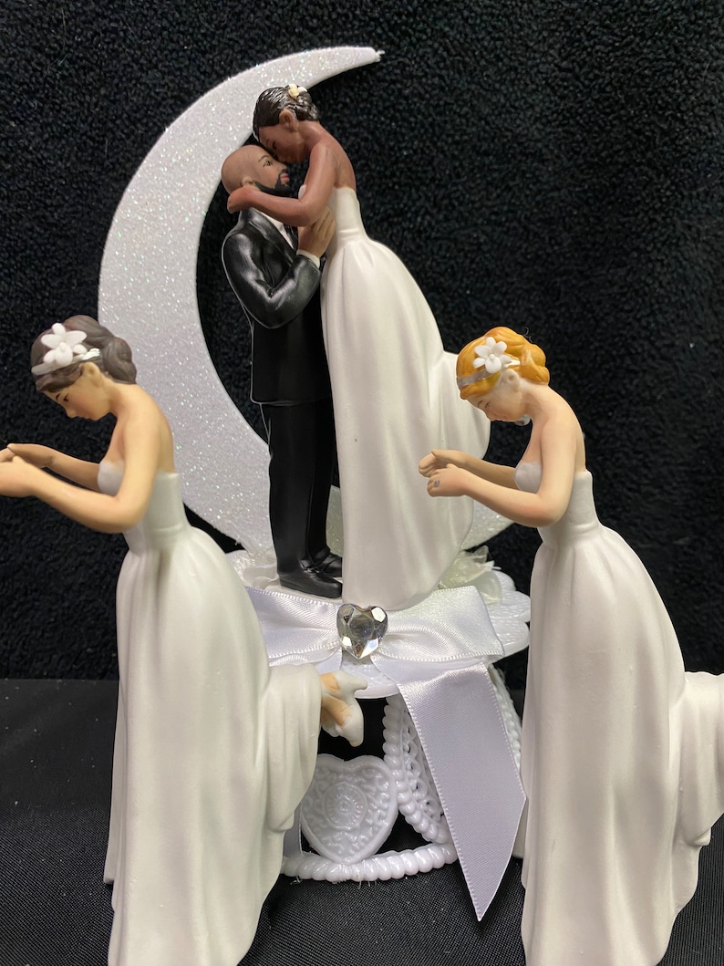 Pu&ograve; includere: Un topper per torta nuziale con una coppia che si bacia sotto una luna crescente, con altre due figure inginocchiate su entrambi i lati. Le figure sono tutte vestite con abiti da sposa bianchi.