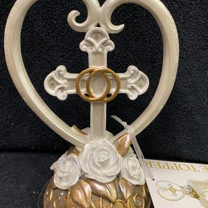 May include: White ceramic cake topper featuring a heart-shaped frame with a gold cross and two intertwined rings. The base is decorated with white roses and gold accents.