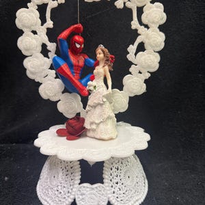 May include: A cake topper featuring a figurine of a superhero in a red and blue suit suspended within a white glittery heart-shaped frame of roses, positioned above a figurine of a person in a white dress, set on a white scalloped pedestal base.