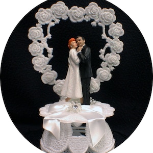 May include: A white cake topper featuring a bride and groom figurine standing in front of a white heart-shaped rose garland. The bride is wearing a white dress with glitter and the groom is wearing a black suit. The cake topper is on a white cake stand with a white ribbon.