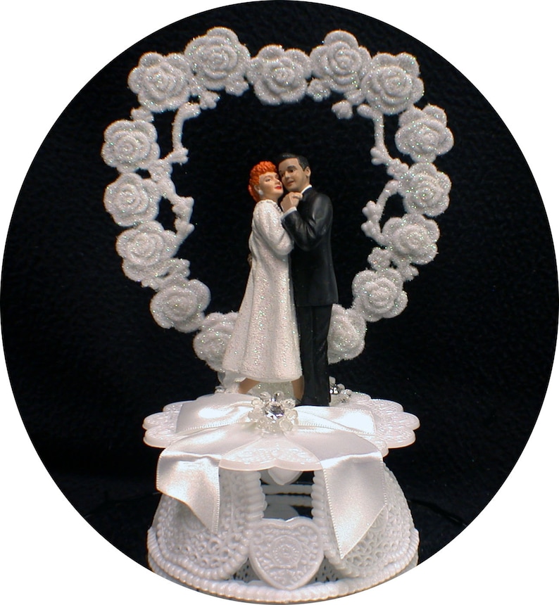 May include: A white cake topper featuring a bride and groom figurine standing in front of a white heart-shaped rose garland. The bride is wearing a white dress with glitter and the groom is wearing a black suit. The cake topper is on a white cake stand with a white ribbon.
